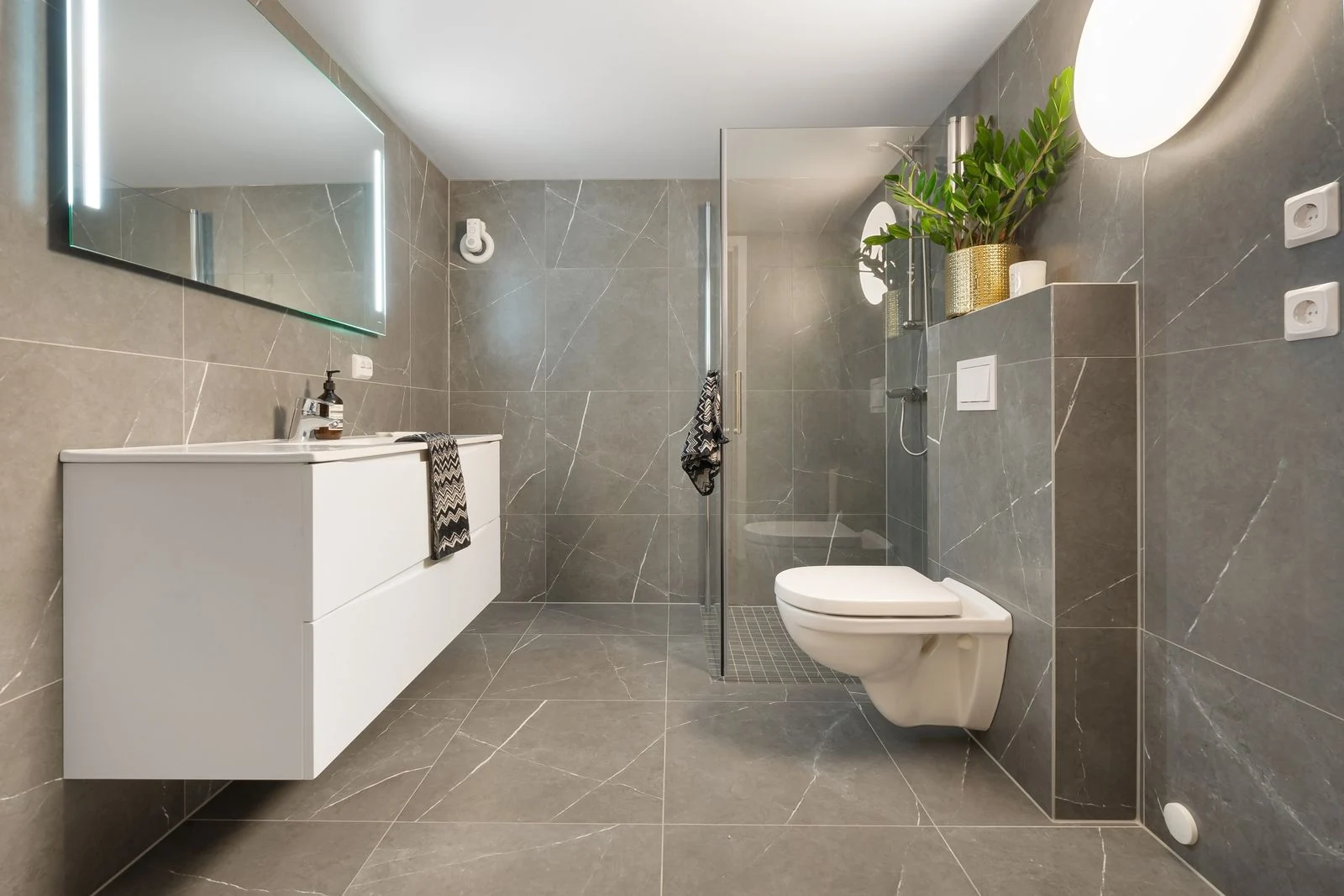 Stylish gray tiled bathroom featuring a shower, toilet, and illuminated mirror